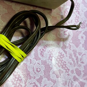 May include: A coiled brown electrical cord secured with a bright yellow band. The end of the cord shows exposed wires. The background is a pink surface with a white floral lace pattern.