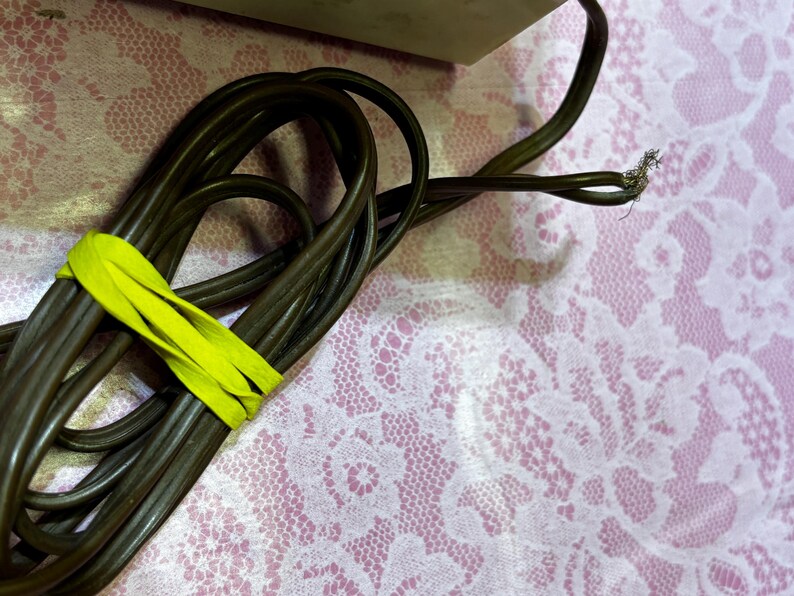 May include: A coiled brown electrical cord secured with a bright yellow band. The end of the cord shows exposed wires. The background is a pink surface with a white floral lace pattern.