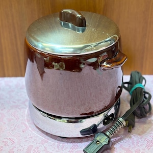 May include: A vintage brown and silver electric slow cooker with a lid and handles. The appliance has a chrome base and a power cord with a plug. The slow cooker is designed for cooking food.