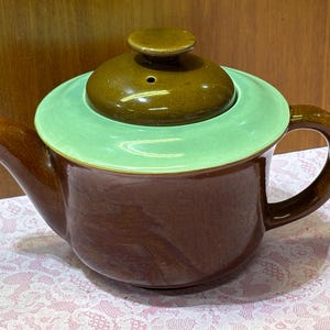 May include: A brown ceramic teapot with a light green rim and lid. The teapot has a curved handle and spout, and the lid has a small knob. The teapot is sitting on a patterned surface.