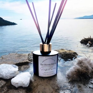 May include: A black reed diffuser with a gold-coloured collar and dark reeds. The label reads "The Hunny Bay Co. Strangford Serenity". The diffuser is placed on a rock near the water, with white rocks and a feather-like object.
