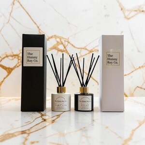 May include: Two reed diffusers with black sticks and gold collars, one in a white bottle and one in a black bottle, each with a label reading The Hunny Bay Co. They are accompanied by a tall black box and a tall white box, both featuring the same brand name.