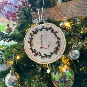 May include: A handmade Christmas bauble featuring a wreath with green leaves and red berries, embroidered with the letter "L" in red and white thread.