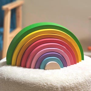 May include: A wooden rainbow stacker toy with seven arched pieces in various colors: green, yellow, orange, pink, purple, light blue, and natural wood. The toy is designed to nest together, creating a rainbow shape. It sits on a white, fluffy surface.