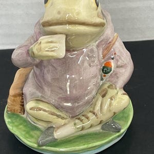 Peut inclure: Figurine en céramique d'une grenouille portant un manteau rose et tenant une tasse. La grenouille est assise sur un nénuphar vert avec une base bleu clair. La figurine a un design détaillé et est peinte avec diverses couleurs.
