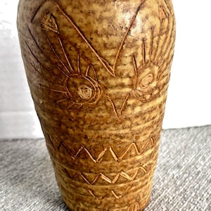 May include: A textured, cylindrical vase in shades of tan and brown. The ceramic vase features etched sun and geometric patterns. The top rim is uneven, and the vase has a rustic, handcrafted appearance. A decorative piece for home decor.