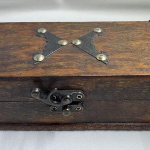 May include: A rectangular wooden box with a dark brown finish. The box has a decorative metal clasp and hinges, and an ornate metal accent on the lid. The box is likely for storage or display.