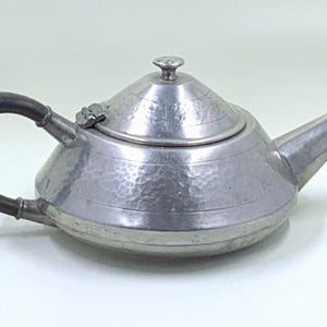 May include: A silver teapot with a hammered finish and a black handle. The teapot has a round body and a domed lid.