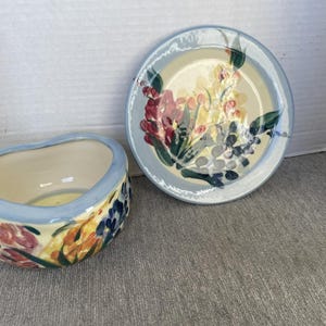May include: A ceramic bowl and a matching plate featuring a hand-painted floral pattern with red, yellow, and green blossoms. Both pieces have a cream-coloured interior and a light blue rim, showcasing a coordinated design with organic shapes.