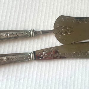 May include: Silver cake server and knife set. The handles feature an ornate floral design, and the blades are engraved with similar patterns. The set is likely vintage, suitable for serving cakes and pastries.
