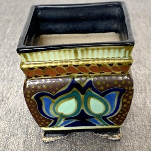 May include: A small, square ceramic planter with a black interior and a decorative exterior. The planter features a brown base with blue and green floral designs, accented with gold and orange details. The planter sits on small feet.