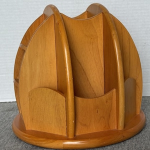 May include: A light brown wooden desk organiser with multiple compartments. The organiser has a rounded base and curved dividers, creating slots for storing items. The wood grain is visible, and the finish is glossy.