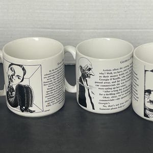 May include: Four white ceramic mugs, each featuring a black and white caricature illustration of a historical figure alongside printed biographical text. The mugs display portraits of Sigmund Freud, Emily Dickinson, Georgia O'Keeffe, and Edgar Allan Poe.