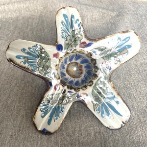 May include: A star-shaped ceramic ashtray with a central well and five points. The ashtray is white with blue, green, and brown floral and leaf designs. The edges are a dark brown colour.