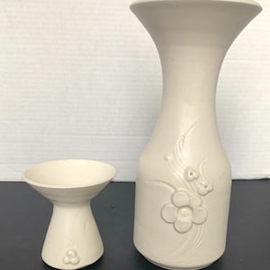 May include: A set of two white ceramic vases with floral designs. The larger vase is taller and has a wider opening than the smaller vase.