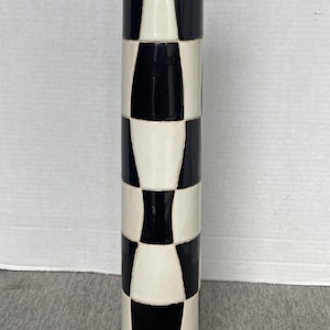 May include: A tall, cylindrical ceramic vase featuring a high-gloss finish with a repeating black and white chequered pattern. The design consists of alternating vertical columns of black and white rectangular shapes arranged in a grid-like formation.