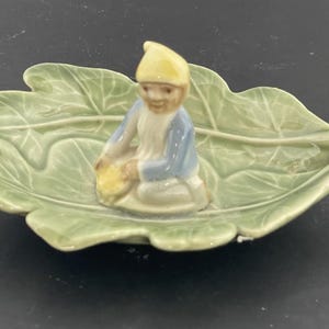 May include: A ceramic gnome figurine kneeling on a large, pale green leaf-shaped dish. The gnome is wearing a yellow hat and blue jacket, holding a yellow basket. The dish has detailed leaf vein patterns.