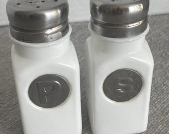 May include: A pair of white salt and pepper shakers with stainless steel lids. The shakers are rectangular with a circular metal emblem on the front, one with the letter "P" and the other with the letter "S".