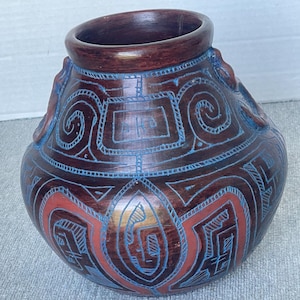 May include: A rounded dark brown ceramic vessel featuring a wide mouth and two small side handles. The surface is decorated with etched geometric patterns highlighted in bright blue and terracotta red, with the letters DKB inscribed on the front.