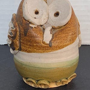 May include: A ceramic owl figurine with brown, white and green glaze. The owl has large, round eyes and a beak. The base of the figurine is decorated with a row of small, rounded shapes.