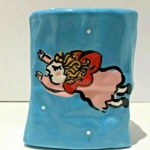 May include: A blue ceramic vase featuring a whimsical illustration of a girl with blonde hair and a red and pink dress flying through the air. The vase has a textured surface and white dots.