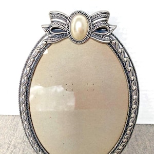 May include: Oval silver-toned picture frame with a bow detail at the top and a faux pearl centre. The frame has an ornate, scrolling design around the edge and a neutral-coloured backing.