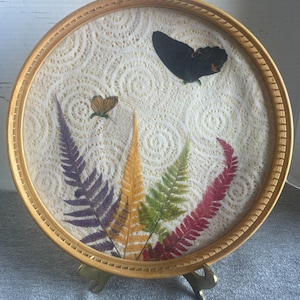 May include: A circular artwork in a light brown wooden frame, displaying a cream-colored, patterned background. The artwork features two butterflies and several colorful fern fronds. The frame is supported by a brass stand.