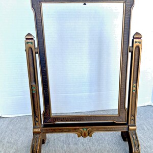 May include: An antique-style vanity mirror with a rectangular frame. The frame is a dark bronze colour with gold accents and colourful details. The mirror has a decorative base and top, and is supported by two vertical posts.