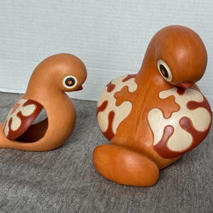 May include: Two ceramic bird ornaments in shades of orange and cream. One bird is seated, the other is a hollowed-out shape. Both have dark eyes and cream-coloured patterns with reddish-brown outlines. The ornaments are decorative.