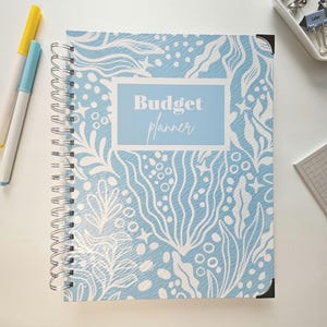May include: A spiral-bound budget planner with a light blue cover featuring a white, abstract floral design. The words "Budget planner" are in a light blue rectangle. Yellow and blue markers are visible.