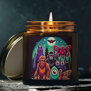 Puede incluir: Una vela encendida en un frasco de vidrio con tapa dorada. La etiqueta presenta una colorida ilustración de varios monstruos de dibujos animados, incluyendo un Bigfoot, un gorila y una polilla. La vela está sobre una superficie oscura.
