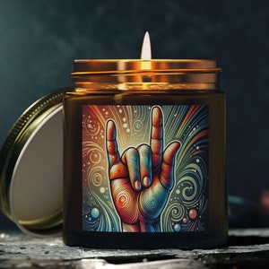 May include: A lit scented candle in a brown glass jar with a gold lid. The label features a colorful illustration of a hand making the rock and roll gesture. The candle sits on a dark surface.