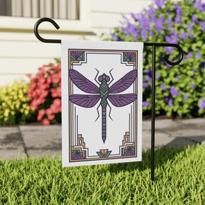 Peut inclure: Un drapeau de jardin décoratif avec un motif de libellule stylisée. La libellule est violette et verte, sur fond blanc avec une bordure d'inspiration Art Déco en or, vert et violet. Le drapeau est affiché sur un support en métal noir dans un jardin.