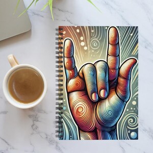 Puede incluir: Una libreta de espiral con una ilustración colorida de una mano haciendo el signo de 'te amo'. El fondo es un patrón de remolinos de azules, verdes y amarillos.