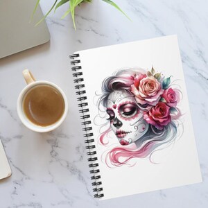May include: A spiral-bound notebook with a watercolour illustration of a sugar skull adorned with pink roses and flowing hair. The notebook is open, showing blank white pages. A coffee cup is on a marble surface.