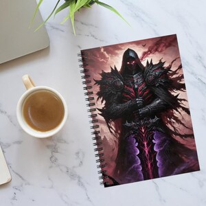 May include: Spiral-bound notebook featuring a fantasy illustration of a dark knight in black armor with red accents. The artwork depicts a hooded figure with glowing red eyes, holding a glowing sword against a cloudy, reddish background. A cup of coffee and a laptop are also visible.