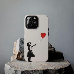 Puede incluir: Una funda blanca para iPhone con una imagen en blanco y negro de una niña que alcanza un globo rojo en forma de corazón. La funda tiene un recorte para el objetivo de la cámara.