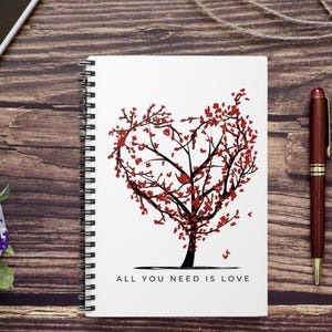 May include: A white spiral bound notebook with a black and red tree illustration in the shape of a heart. The tree has red leaves and the text "ALL YOU NEED IS LOVE" is printed below the tree.