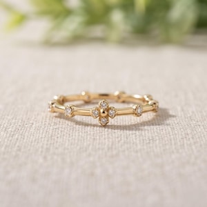 Puede incluir: Un anillo fino de oro con un patrón repetitivo de pequeñas piedras redondas y transparentes engastadas en la banda. El centro del diseño muestra un motivo de flor de cuatro pétalos formado por cuatro piedras alrededor de una cuenta de oro.