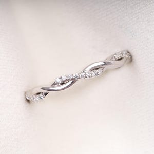 Pode incluir: Um anel prateado com um design torcido, adornado com pequenas pedras preciosas brilhantes. A banda apresenta um padrão entrelaçado, criando uma aparência delicada e elegante. As pedras preciosas são espaçadas uniformemente ao longo da superfície do anel.