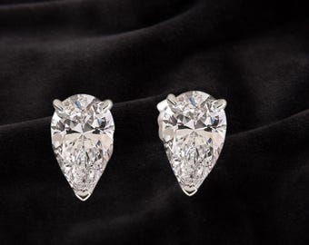 Può includere: Un paio di orecchini a lobo con diamanti a forma di pera scintillanti. Le pietre preziose chiare e sfaccettate sono incastonate in una montatura in argento o oro bianco, presentate su sfondo nero. Gli orecchini sono progettati per catturare la luce.