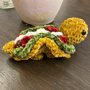 May include: A crocheted plush toy shaped like a turtle with a taco shell on its back. The taco shell is made of green, yellow, and red yarn. The turtle is made of yellow yarn.