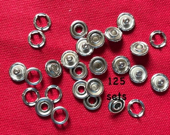 May include: A close-up of 125 sets of silver metal snap fasteners. The fasteners are arranged in a random pattern on a red fabric surface.