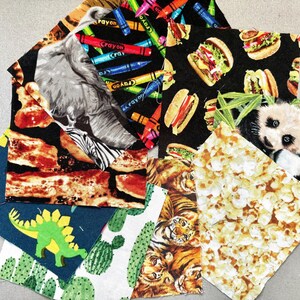May include: A collection of assorted fabric squares featuring various printed patterns including colorful crayons, hamburgers, a zebra, a panda, popcorn, tigers, cacti, and a green dinosaur on a dark blue background, all arranged in a scattered overlapping pile.