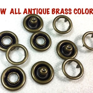 May include: Antique brass color snap fasteners.  There are 9 snap fasteners in the image.  The fasteners are round and have a raised center.  The fasteners are arranged in a random pattern on a white surface.