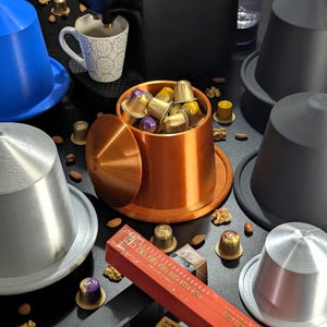 Può includere: Un portacapsule di caffè color rame con un coperchio, pieno di capsule di caffè di diversi tipi. Il portacapsule è su una superficie nera con capsule di caffè sparse intorno. C'è una scatola di capsule di caffè nell'angolo in basso a destra dell'immagine.