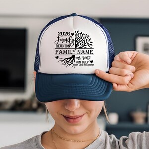 A person is holding up a blue and white baseball cap with a family name and a tree design on it.