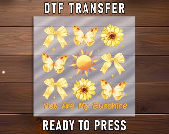 May include: A DTF transfer sheet featuring a grid of yellow graphics including bows, butterflies, sunflowers, and a central sun. The text "You Are My Sunshine" is printed at the bottom. The sheet is labelled "DTF TRANSFER" at the top and "READY TO PRESS" at the bottom.