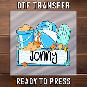 May include: DTF transfer design featuring beach-themed illustrations. Includes a blue bucket with sand, a beach ball character, a popsicle, and a sandcastle. The name "Jonny" is displayed on a sign. The words "DTF TRANSFER" and "READY TO PRESS" are also visible.