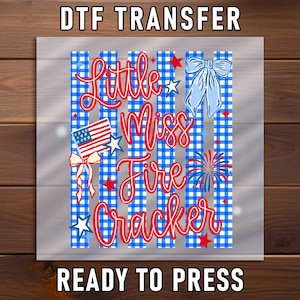 May include: A DTF transfer sheet featuring the text Little Miss Fire Cracker in red script. The design includes a blue and white gingham striped background, a small American flag, a light blue bow, and red and blue star accents.